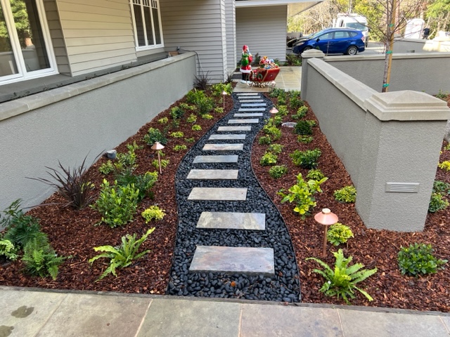 Modern front yard with a curved stone pathway set in black river rocks, surrounded by neatly arranged mulch, green plants, and copper landscape lights, leading to a festive Christmas decoration by the driveway.