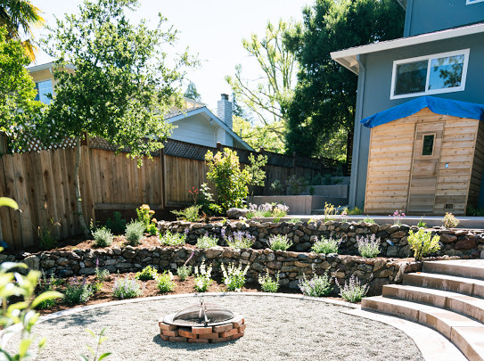 Backyard with terraced stone retaining walls, young plants and flowers, a gravel patio with a brick fire pit in the center, curved stone steps, and a modern shed with a wooden door.
