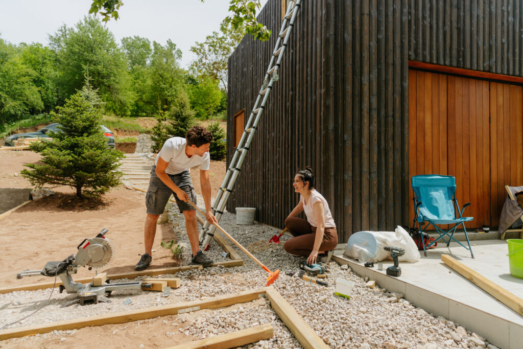 Man and woman working together on an outdoor DIY project, leveling gravel and framing wood near a modern dark wood house with large wooden doors and a ladder leaning against the building.