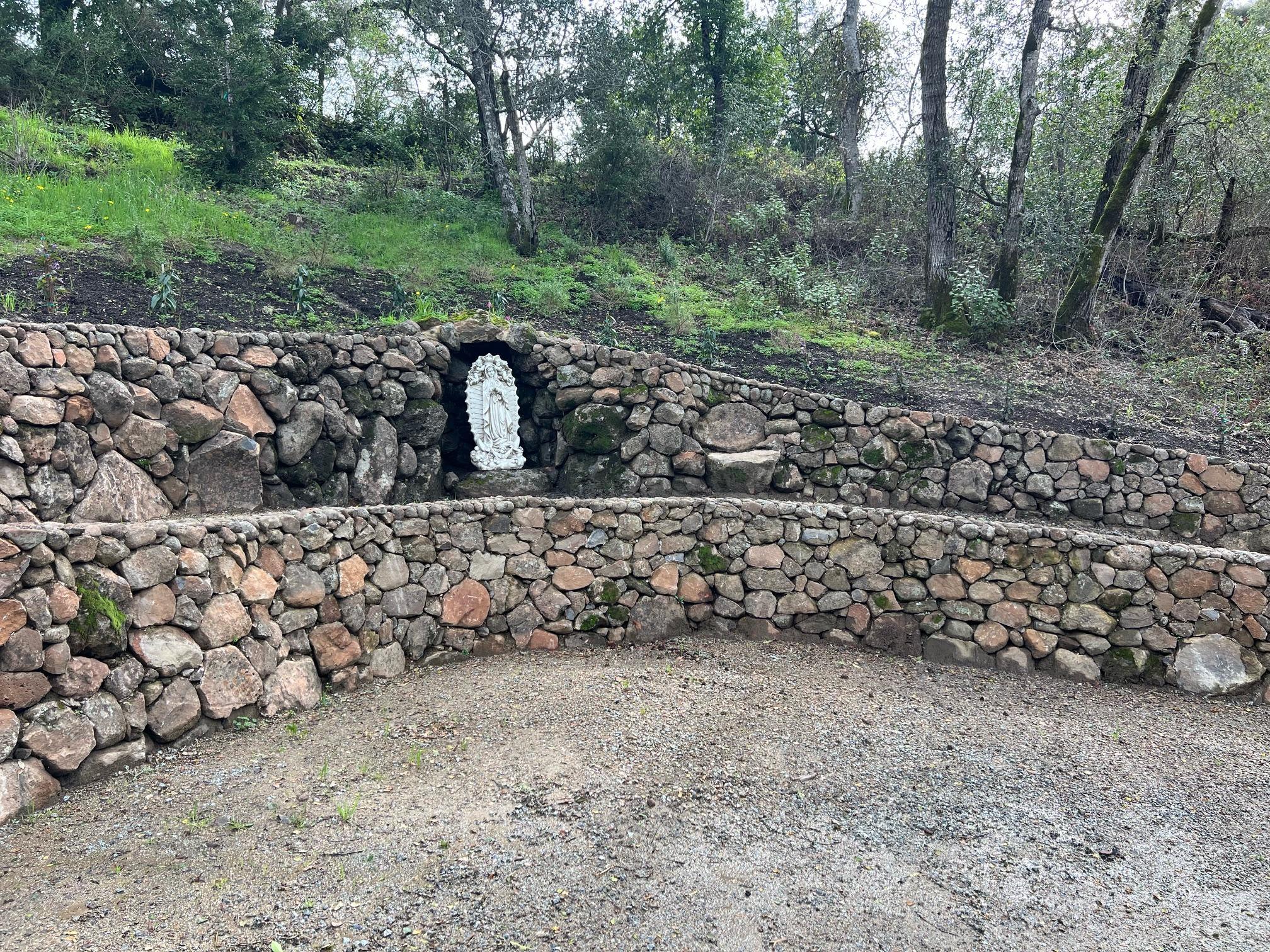 A curved, multi-level retaining wall made of stacked natural stones with a small statue set into a niche.