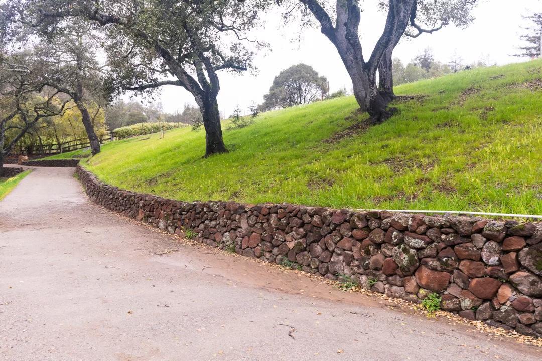 A rustic roadside retaining wall built from stacked natural stones lining a sloped grassy hillside with trees.